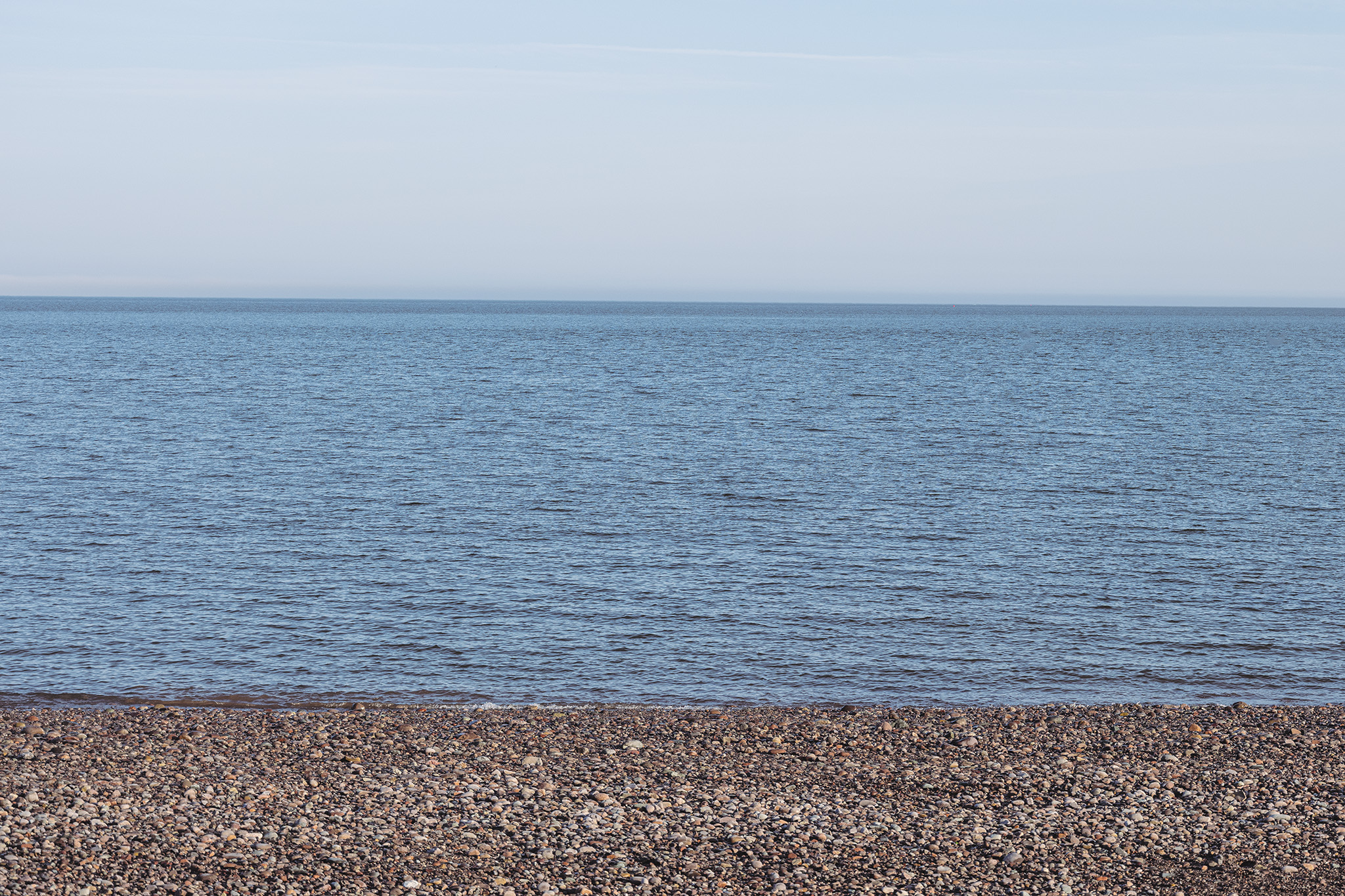 A formal, eye-level study that engages with the strict composition of traditional landscape photography to evoke a sense of quiet isolationism. The frame is divided into three distinct, unyielding horizontal bands: a vast shoreline of multi-colored pebbles in the foreground, a wide expanse of deep blue water in the center, and a pale, cloudless sky above. By deliberately removing any human presence, structures, or tracking markers, the image emphasizes an immense scale and geographic detachment. The unreachable, flat horizon line reinforces a psychological separation, transforming a straightforward coastal view into a stark meditation on distance and solitude.