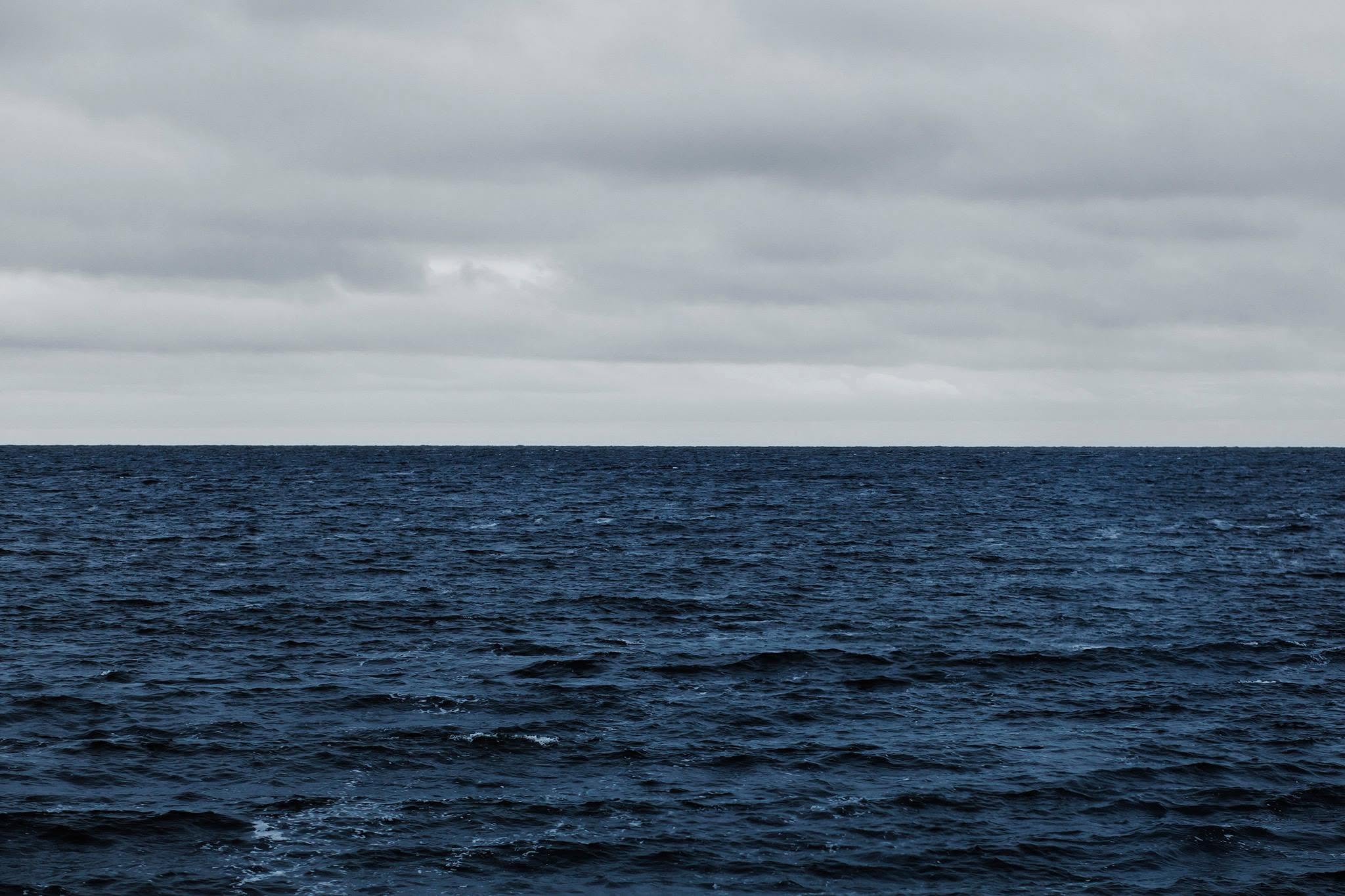 An immense expanse of deep, shadowed indigo water stretches toward a perfectly straight, razor-sharp horizon line that bisects the frame. Above, a heavy, uniform blanket of slate-grey clouds hangs low, casting an oppressive, diffused light that drains the scene of warmth. The surface of the Bay of Fundy is restless, textured with tightly packed, choppy ridges and white crests that evoke a profound sense of isolation, vastness, and a somber, melancholic reverence.The composition captures a stark, minimalist dialogue between sea and sky, channeling the haunting sorrow of Ron Hynes' "Atlantic Blue." Through its cool, restricted palette and tense stillness, the image stands as a quiet, elegiac monument to the souls lost to the depths, reflecting both the sublime beauty and the indifferent, tragic power of the North Atlantic.