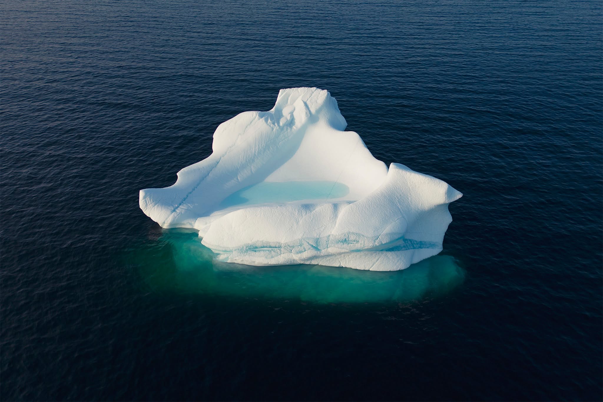 An aerial, bird's-eye perspective captures a solitary, pristine white iceberg floating in the vast expanse of a deep navy blue ocean. The iceberg is sculpted with fine, ridge-like contours along its slopes, featuring a naturally formed hollow at its center that holds a pool of calm, turquoise-meltwater. Extending just beneath the surface of the dark ocean, a luminous, wide shelf of glacial ice glows intensely in vibrant shades of aquamarine and emerald green. The sharp, high-contrast light accentuates the deep shadows of the icy topography, emphasizing themes of primordial geology, sublime isolation, and the fragile light of the North Atlantic coast.