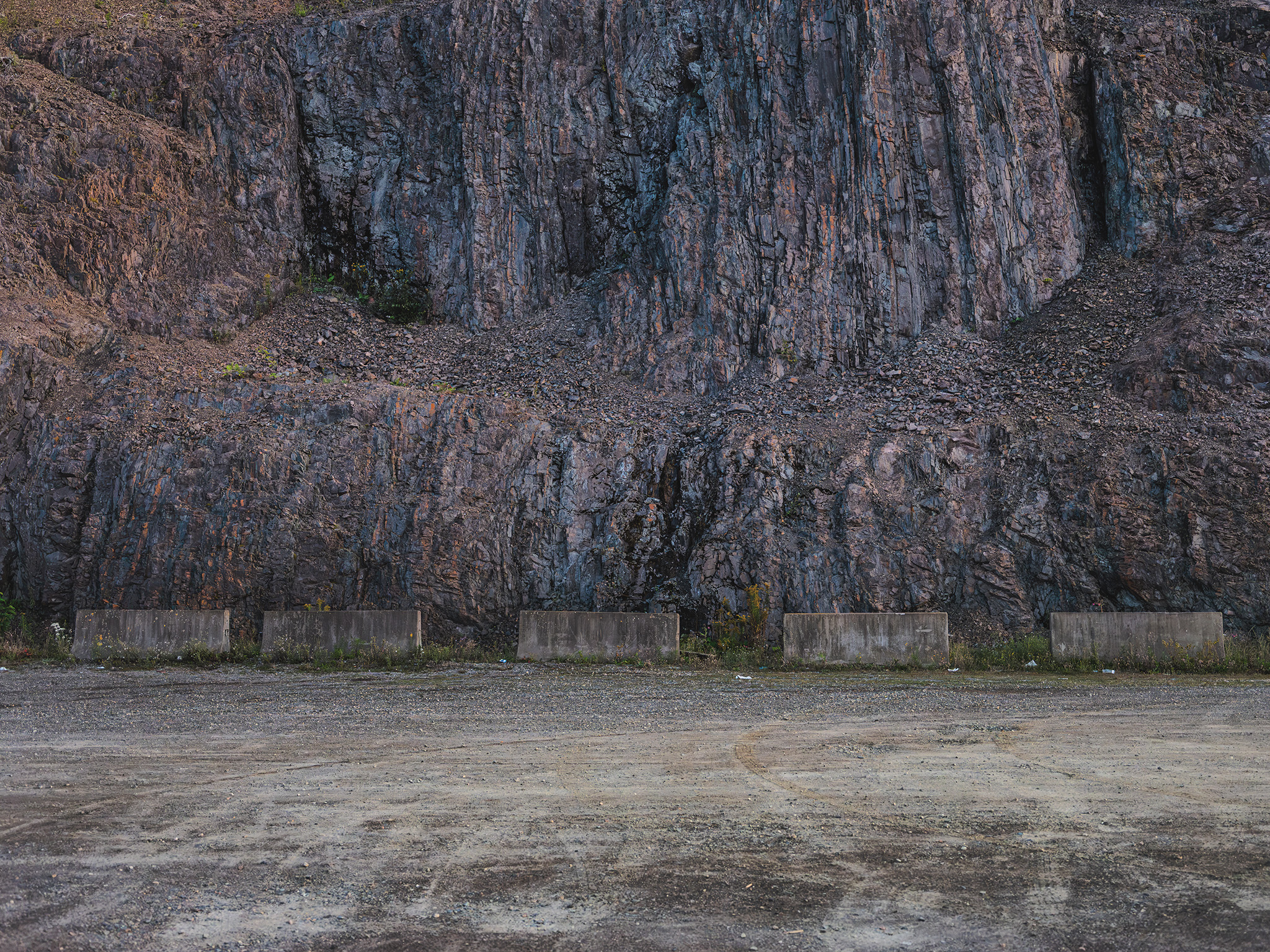 A brutalist study in spatial friction. The work confronts the viewer with a sheared vertical topography—a raw index of geological time violently disrupted by industrial intervention. By positioning a rhythmic line of modular concrete barriers against the chaotic, monumental mass of the rock face, the image interrogates the boundary between deep time and human containment. The stark, gravel foreground serves to flatten the perspective, transforming a scarred landscape into an austere, geometric abstraction on permanence and anthropocentric control.