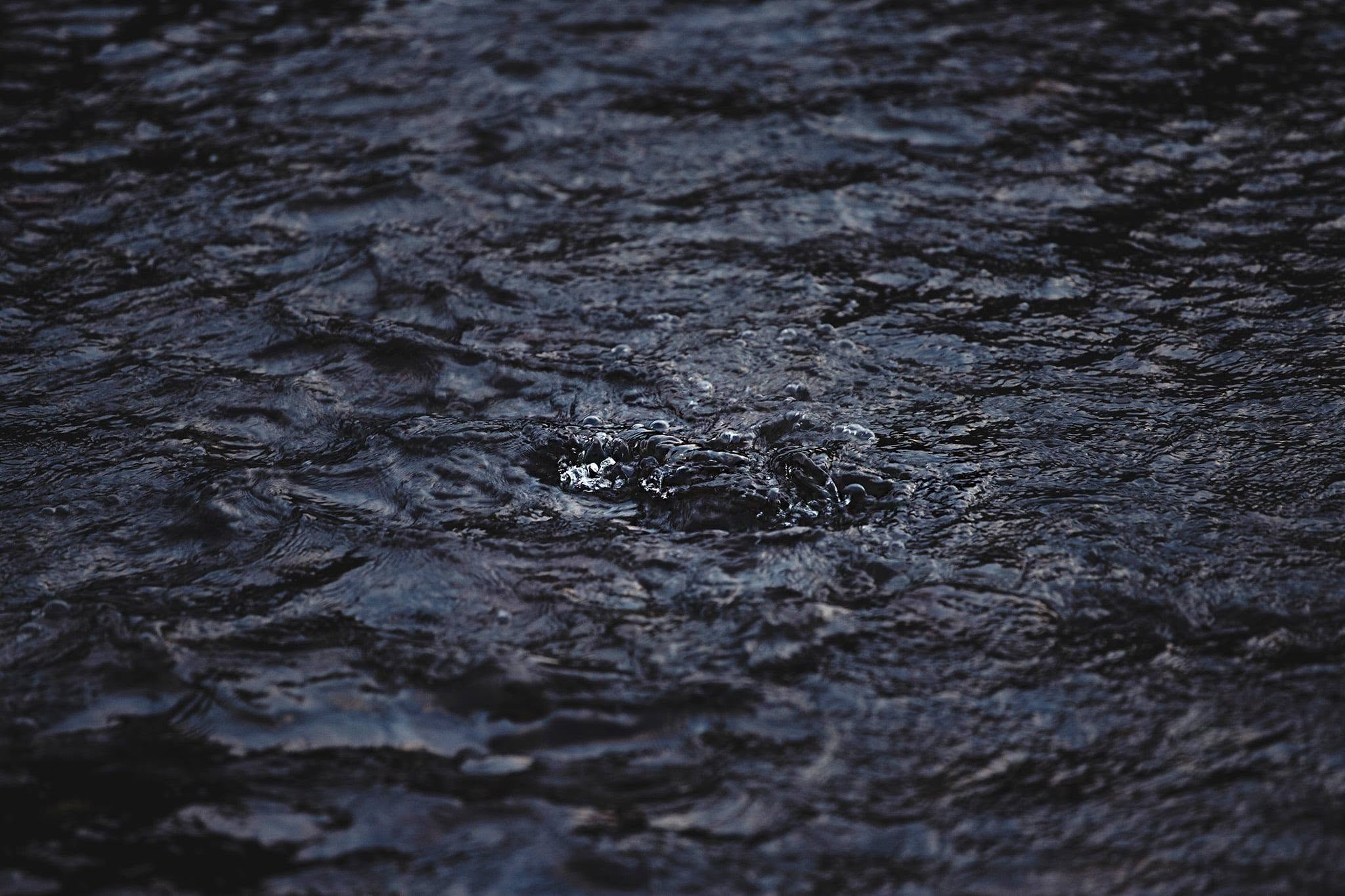  close-up, high-contrast shot captures a dynamic texture of churning water, rendered in a deeply saturated, low-key palette of obsidian, indigo, and slate gray.The composition is heavily planar and fluid, rejecting a traditional horizon line to immerse the viewer entirely in the water's surface tension. At the center of the frame, a localized disturbance breaks the rhythm of the undulating ripples, creating a cluster of crystalline bubbles and churning foam. This central aperture catches the ambient light, introducing sharp, specular highlights that gleam like shattered glass against the darker, velvety surrounding currents.