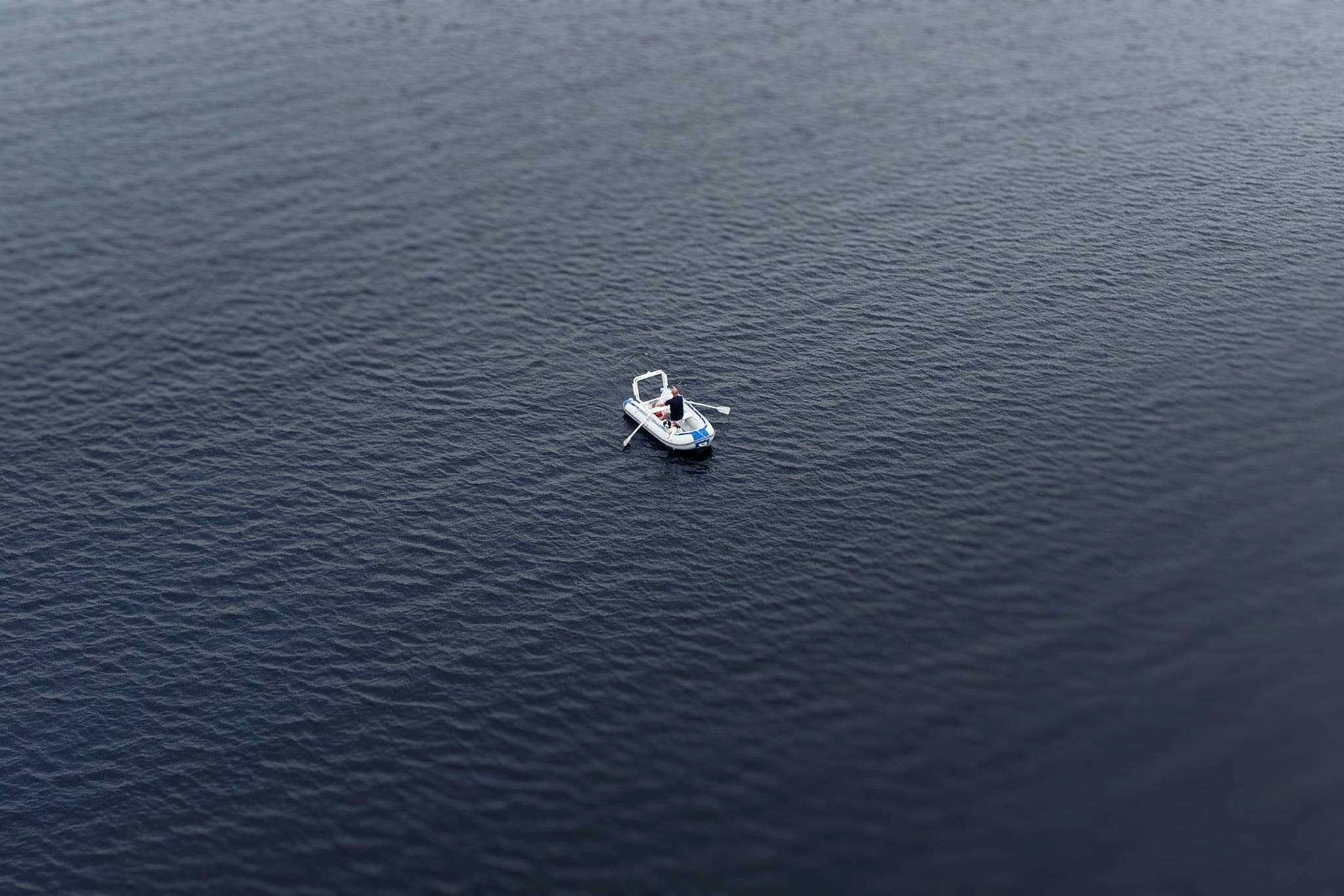 A high-angle, minimalist composition centers a small, white-and-blue inflatable dinghy within an immense, enveloping field of deep charcoal water. A single figure sits inside, oars extended, rendered minute and vulnerable against the vastness. The surrounding water is finely textured with subtle, rhythmic ripples that blur toward the top and bottom of the frame, creating a tilt-shift effect that isolates the vessel in a shallow plane of sharp focus.Serving as the definitive conclusion to the series, the image shifts from the stark abstraction of the open sea to a poignant narrative of human presence and survival. The restricted, monochromatic palette and dramatic scale contrast emphasize themes of isolation, quiet endurance, and the fragile, solitary journey of navigating life on this land and water.