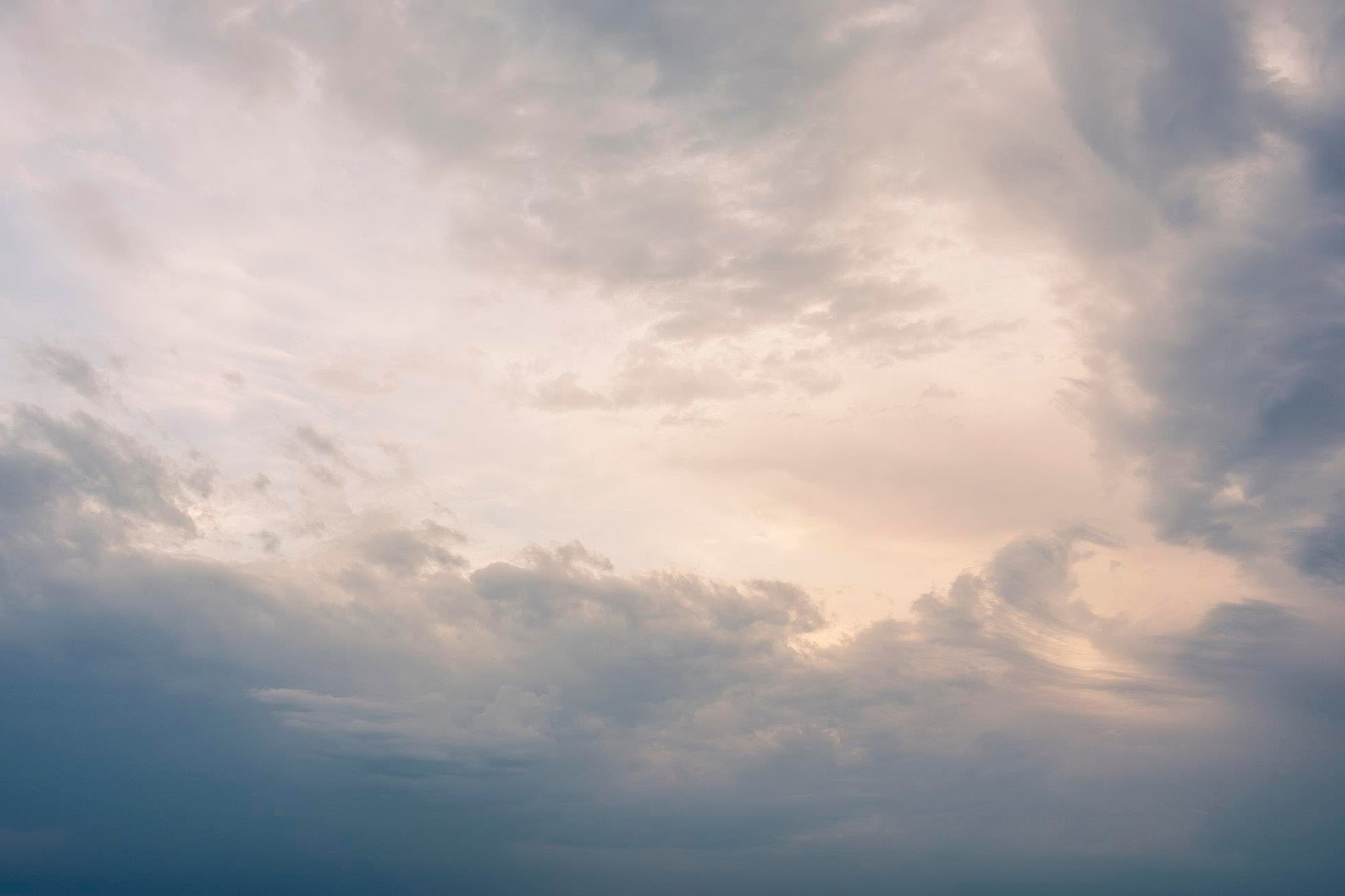 A centralized clearing of soft, luminescent peach and warm ivory light breaks through a heavy framing of muted, slate-blue and stormy grey clouds. 