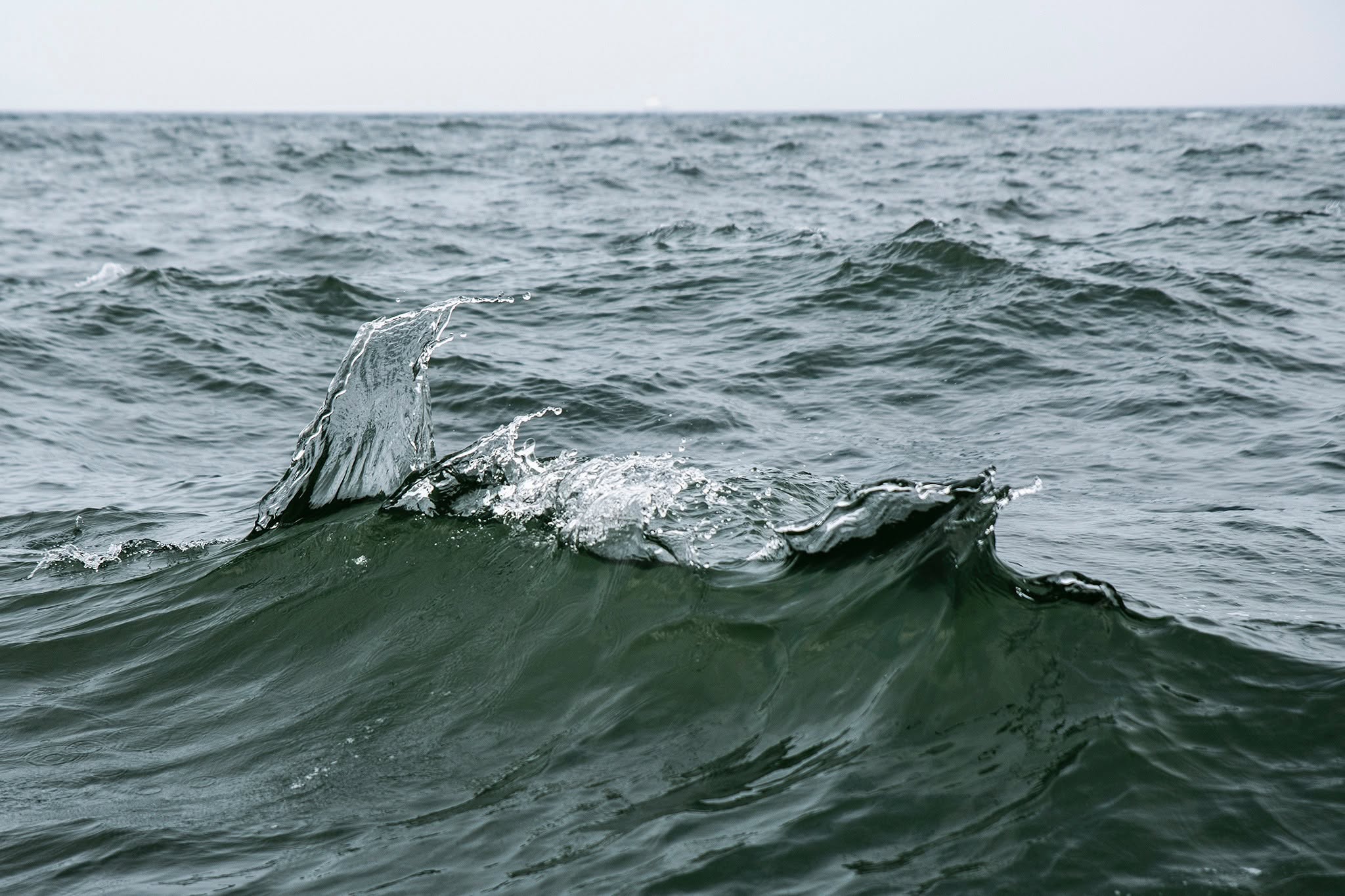 The foreground is dominated by a dark, olive-green wave that swells and crests toward the left. At the apex of this swell, a translucent shard of water fractures upward, echoing the geometry of a dorsal fin. This fluid form is frozen in a high-shutter moment, rendered with crystalline clarity that emphasizes the tension between the liquid medium and atmospheric pressure.